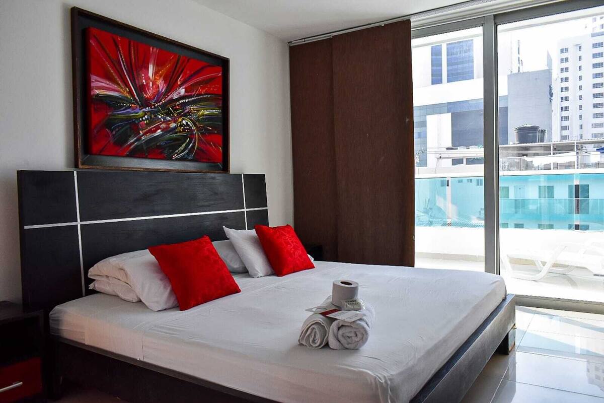 Une chambre d’hôtel avec un grand lit, des oreillers rouges et blancs, un tableau au-dessus du lit et une vue sur les immeubles par la fenêtre.