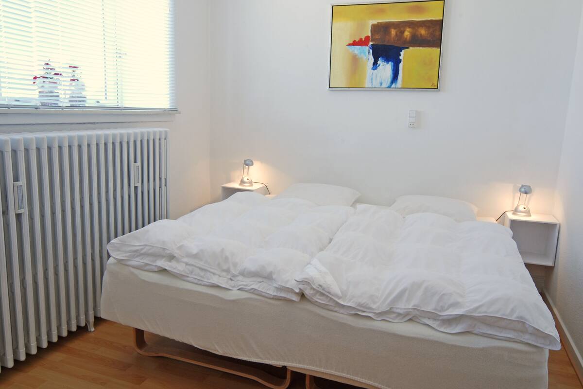 A neatly made bed with white bedding, two bedside lamps, a radiator, and a painting on the wall.