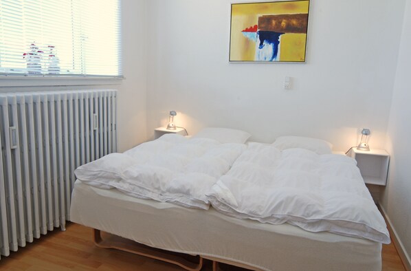 A neatly made bed with white bedding, two bedside lamps, a radiator, and a painting on the wall.