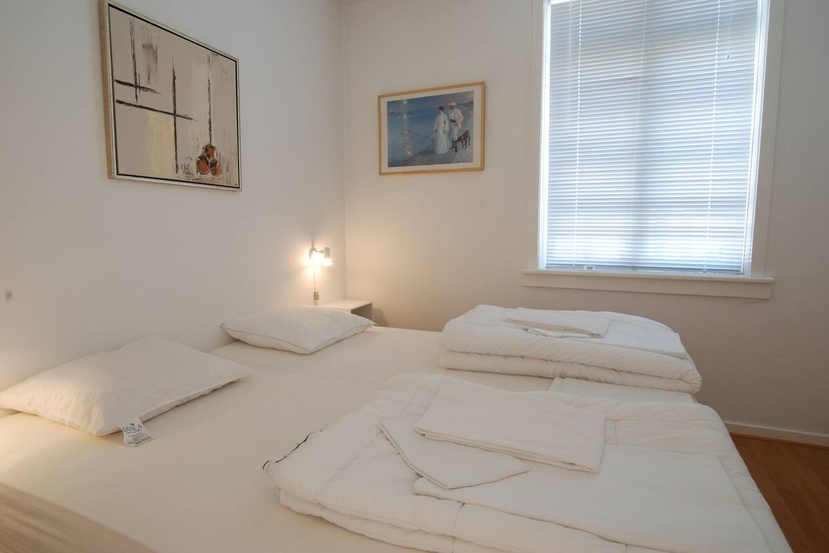 A bedroom with two beds, white bedding, a painting, and a window with blinds.