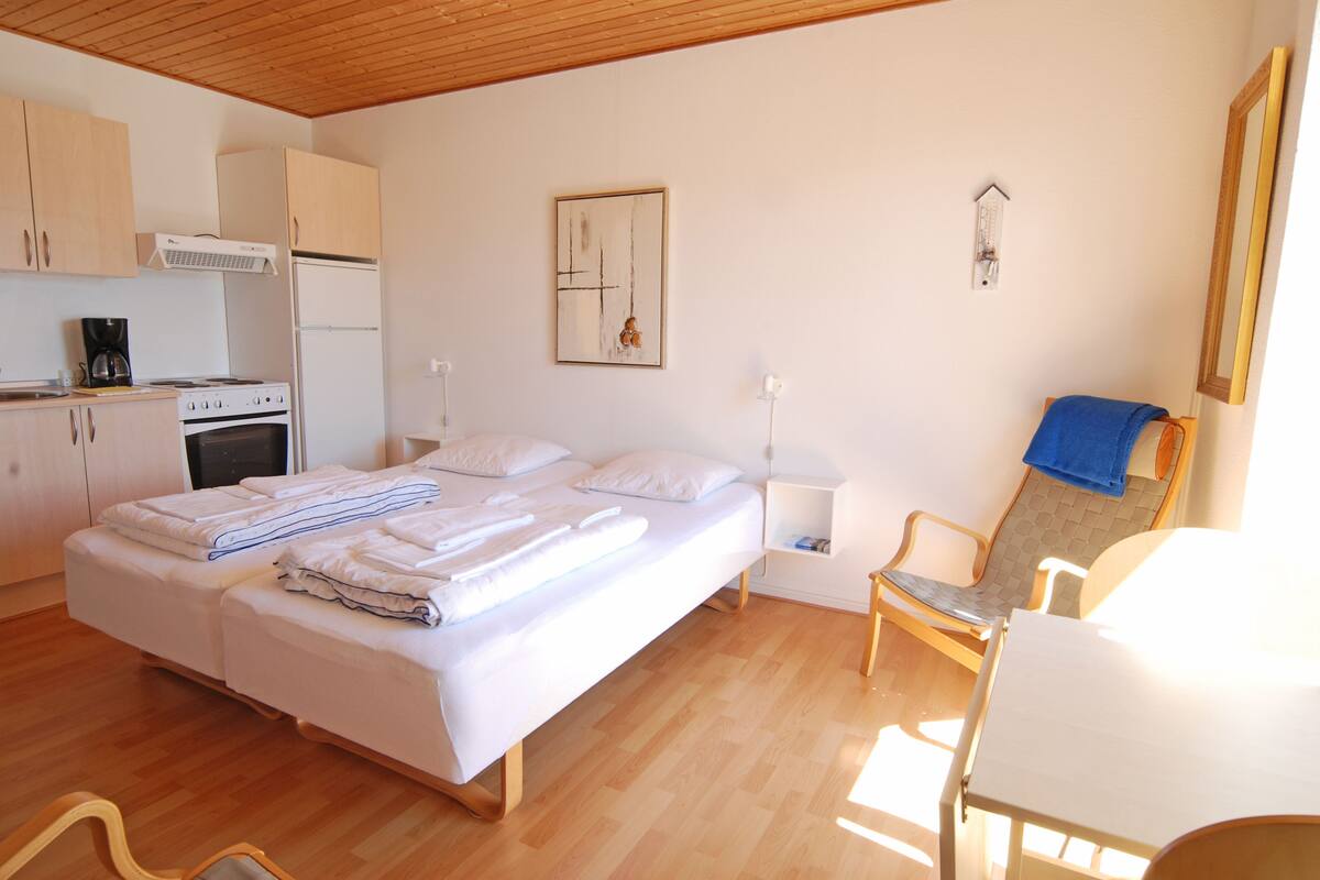A bedroom with a bed, a desk, a chair, a small kitchenette, and a wall-mounted clock.