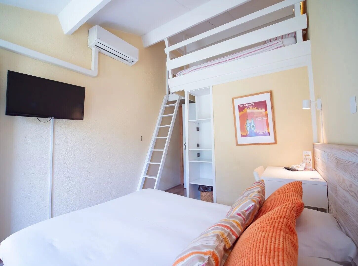 Un dormitorio con una cama litera, un televisor instalado en la pared, una escalera, un cartel enmarcado y una mesita de noche con lámpara.