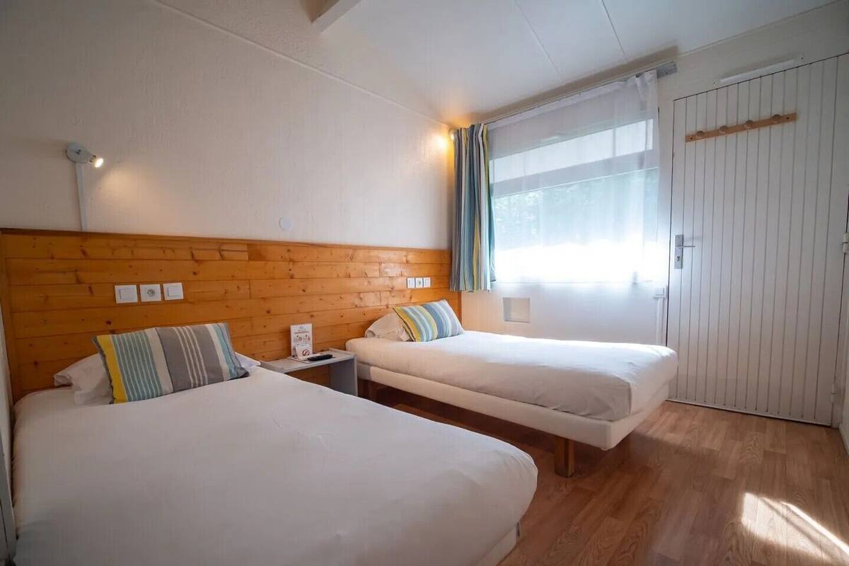 Habitación de hotel con dos camas, cabecero de madera, una ventana con cortinas, un armario y una mesita de noche con lámpara.