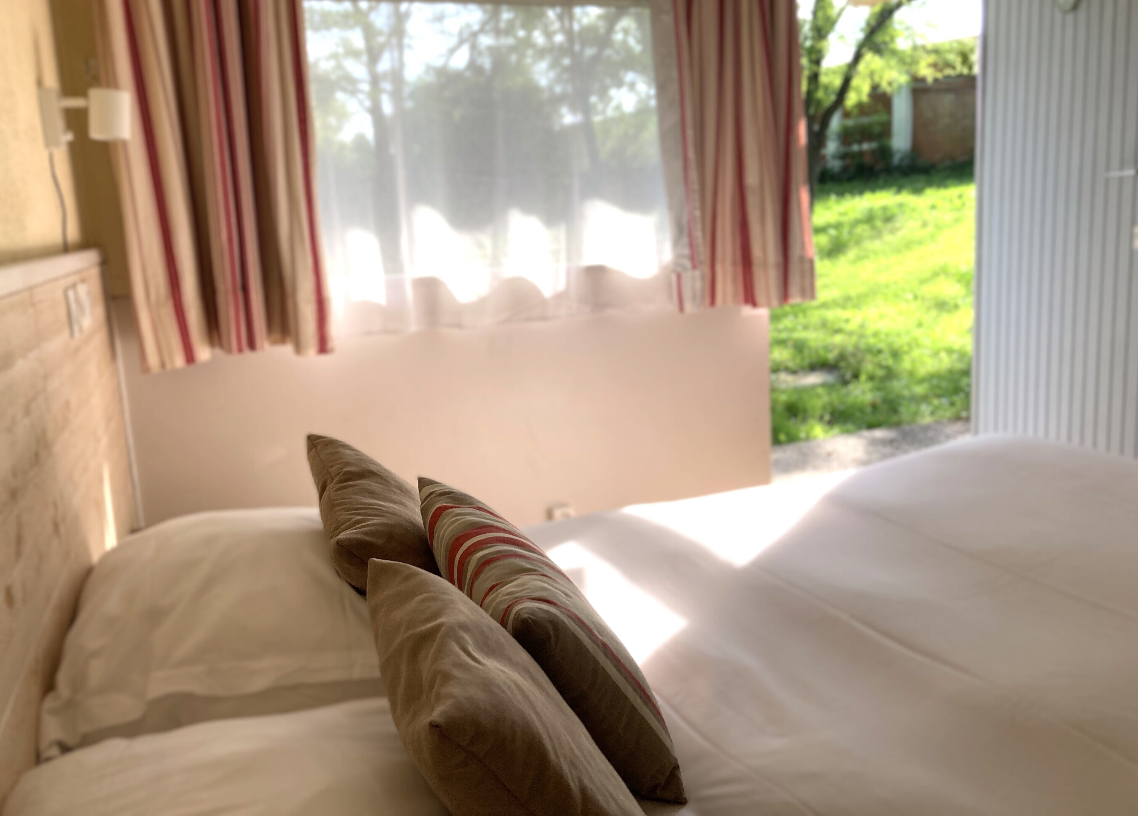 A bed with white linens and two pillows, one striped and one solid, in a room with a window overlooking a green outdoor area.