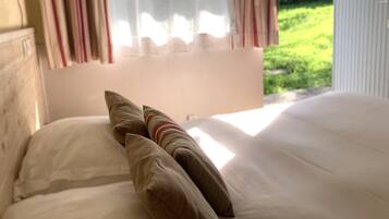 A bed with white linens and two pillows, one striped and one solid, in a room with a window overlooking a green outdoor area.