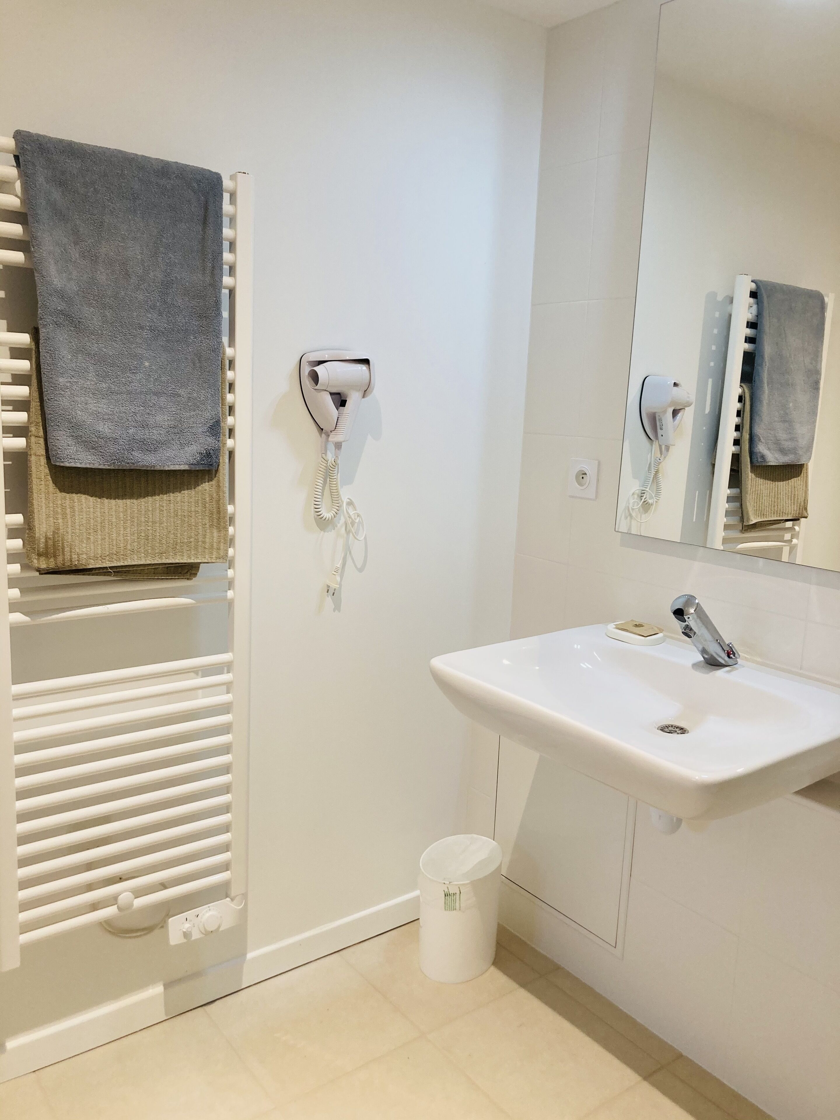 A modern bathroom with a white sink, a towel rack with a grey towel, a hairdryer, and a trash bin.
