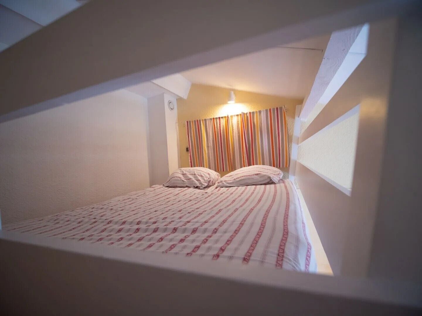 A small, single-bed room with a striped curtain, a visible light switch, and a window on the right.