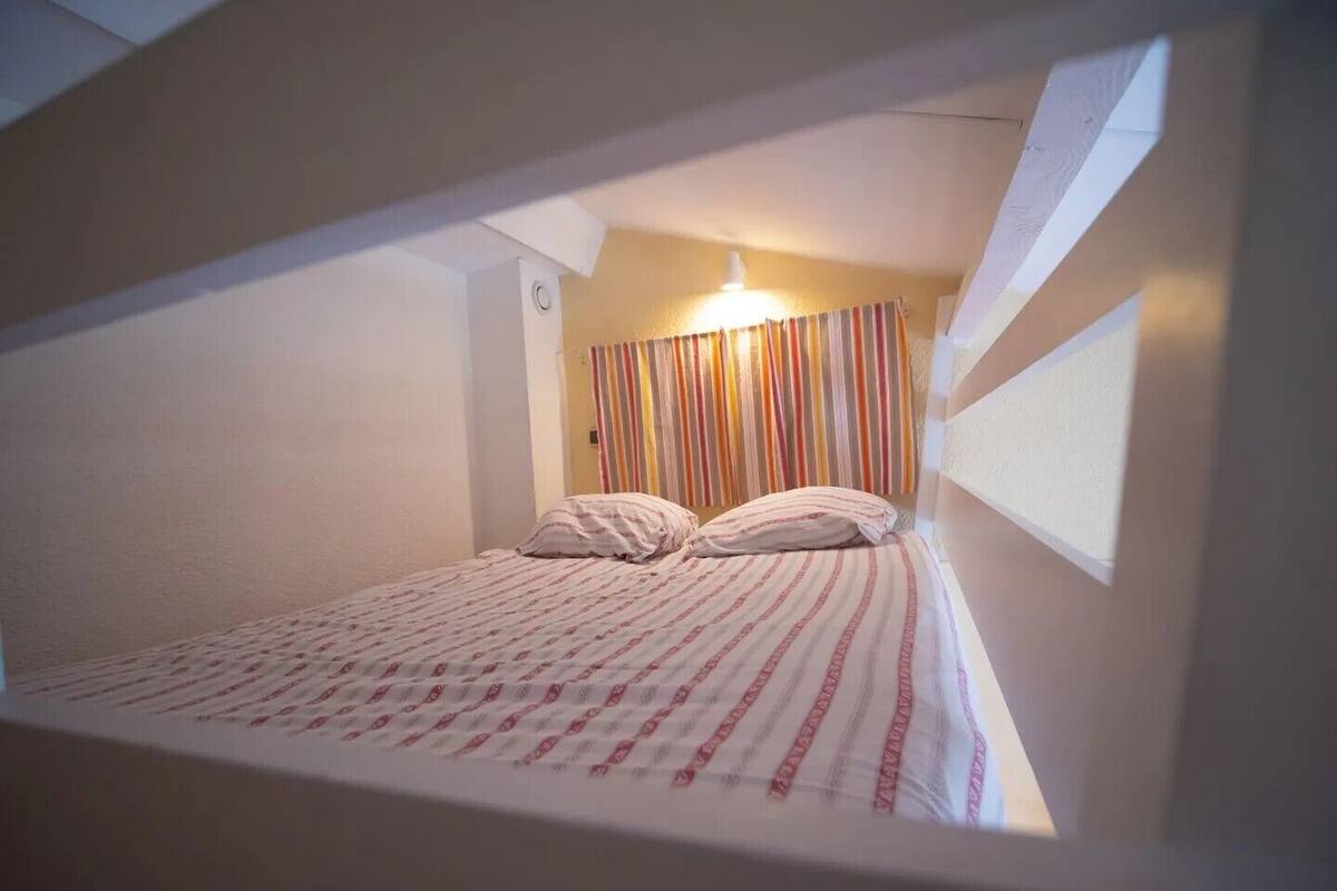 Una habitación pequeña con cama individual, una cortina a rayas, un interruptor de luz visible y una ventana a la derecha.