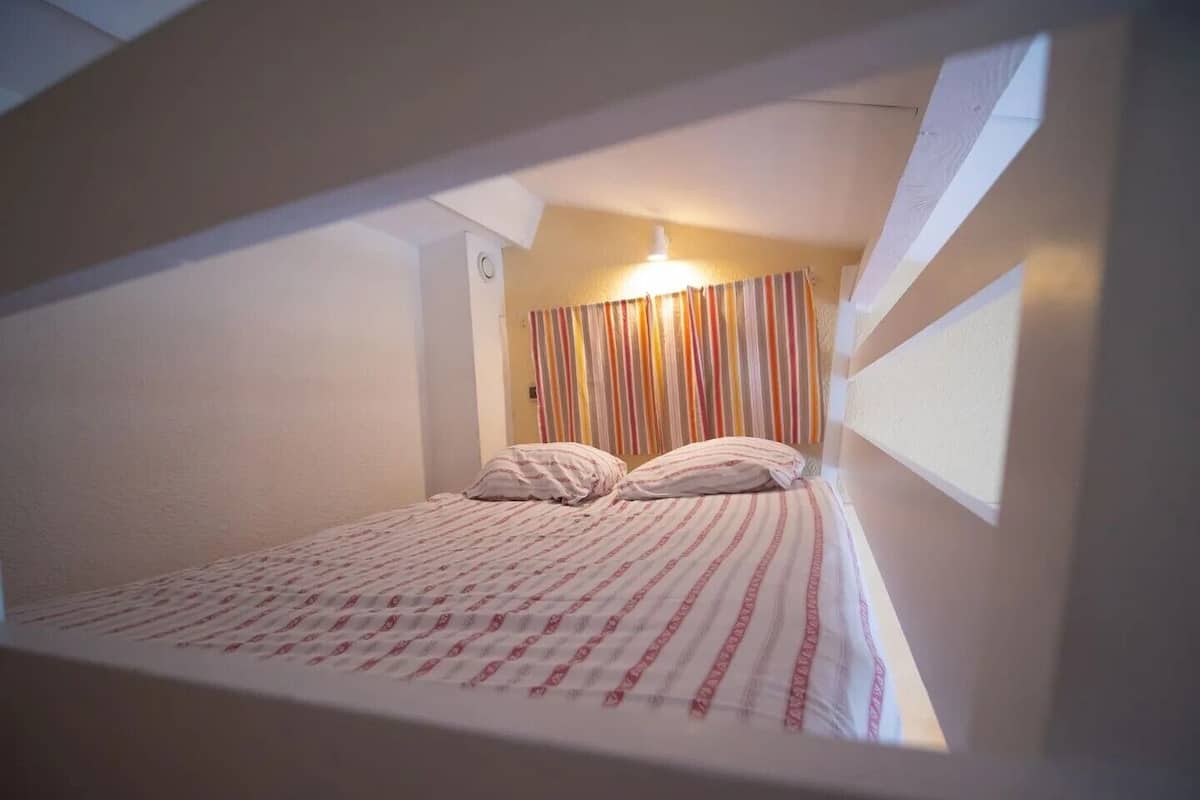 A small, single-bed room with a striped curtain, a visible light switch, and a window on the right.