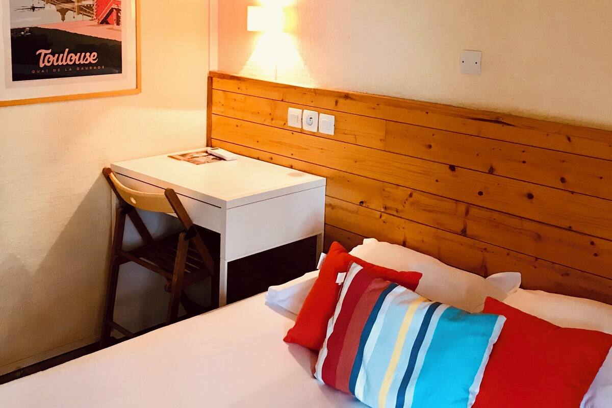Una habitación de hotel con cabecera de madera, una cama individual con almohadas de colores, un escritorio pequeño con una silla y un cuadro en la pared.