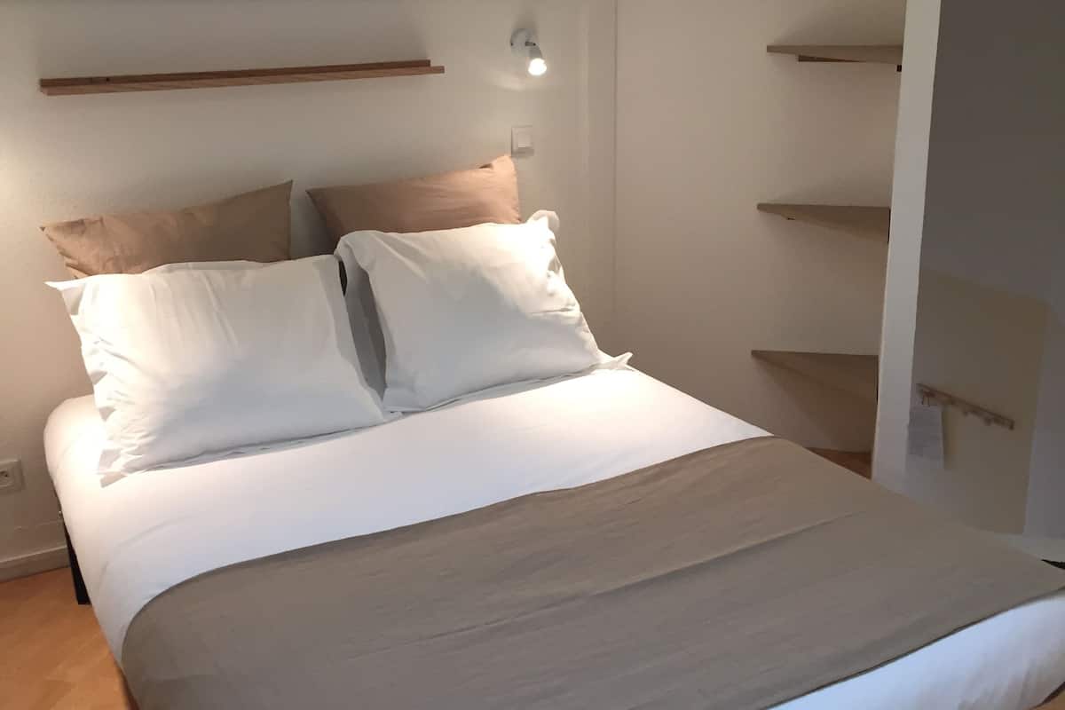 Una cama bien tendida con ropa de cama blanca y color crema, un estante de madera encima y una lámpara fijada a la pared.