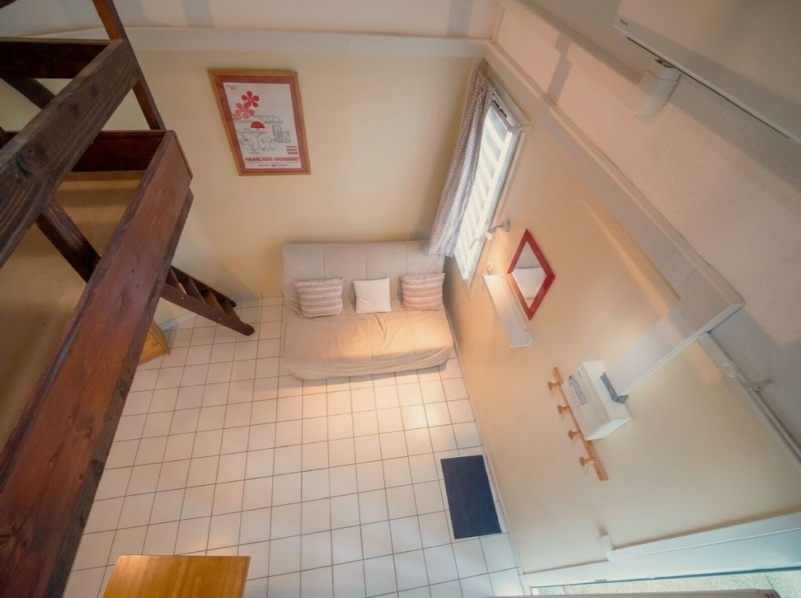 A room with a tiled floor, a bed with pillows, a window with curtains, a wooden staircase, and a framed picture on the wall.