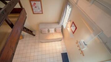 A room with a tiled floor, a bed with pillows, a window with curtains, a wooden staircase, and a framed picture on the wall.