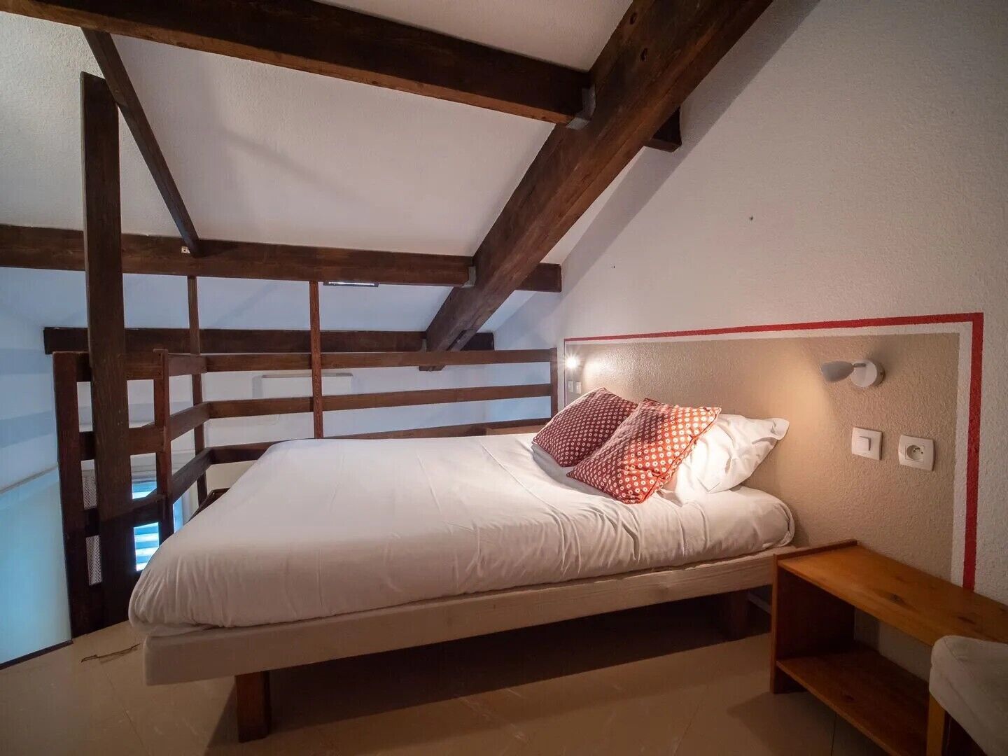 A bedroom with a bed, pillows, a nightstand, a wooden bench, and a view of the sky.