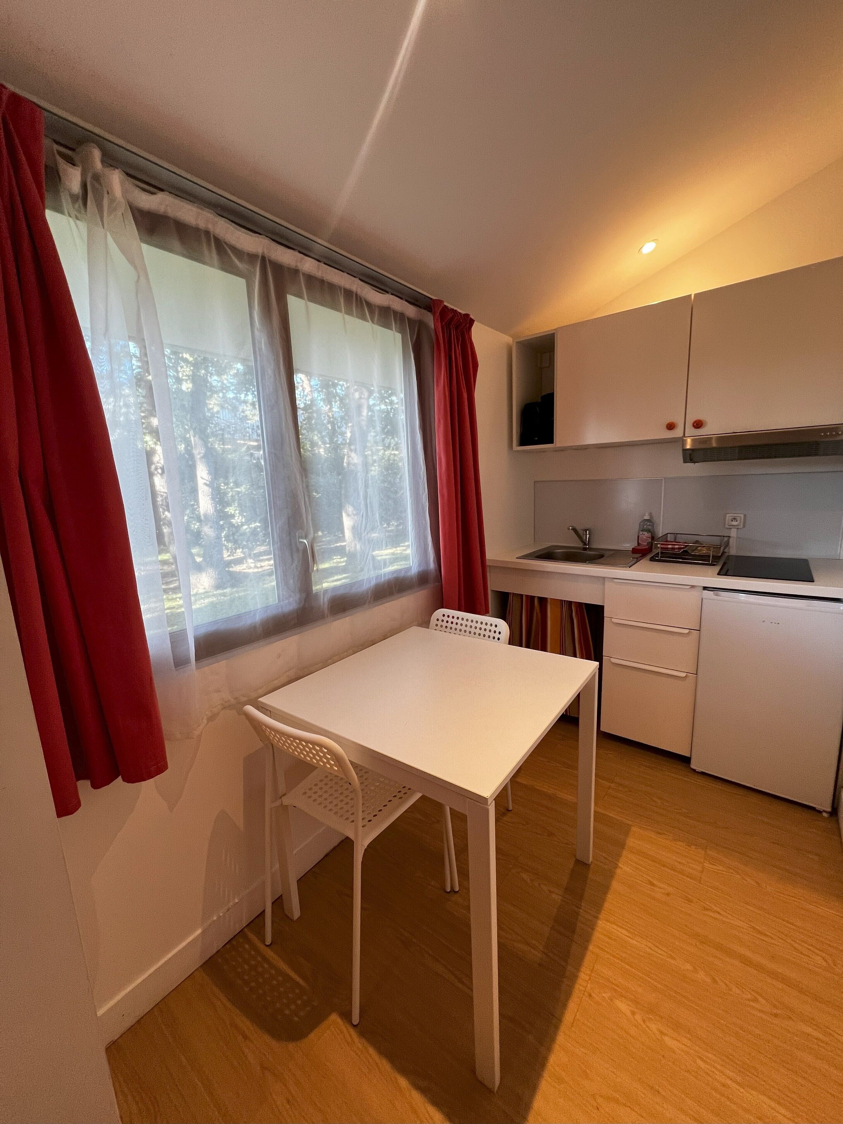 Een kleine, schone kamer met een witte tafel en stoel, een kitchenette en een raam met gordijnen.