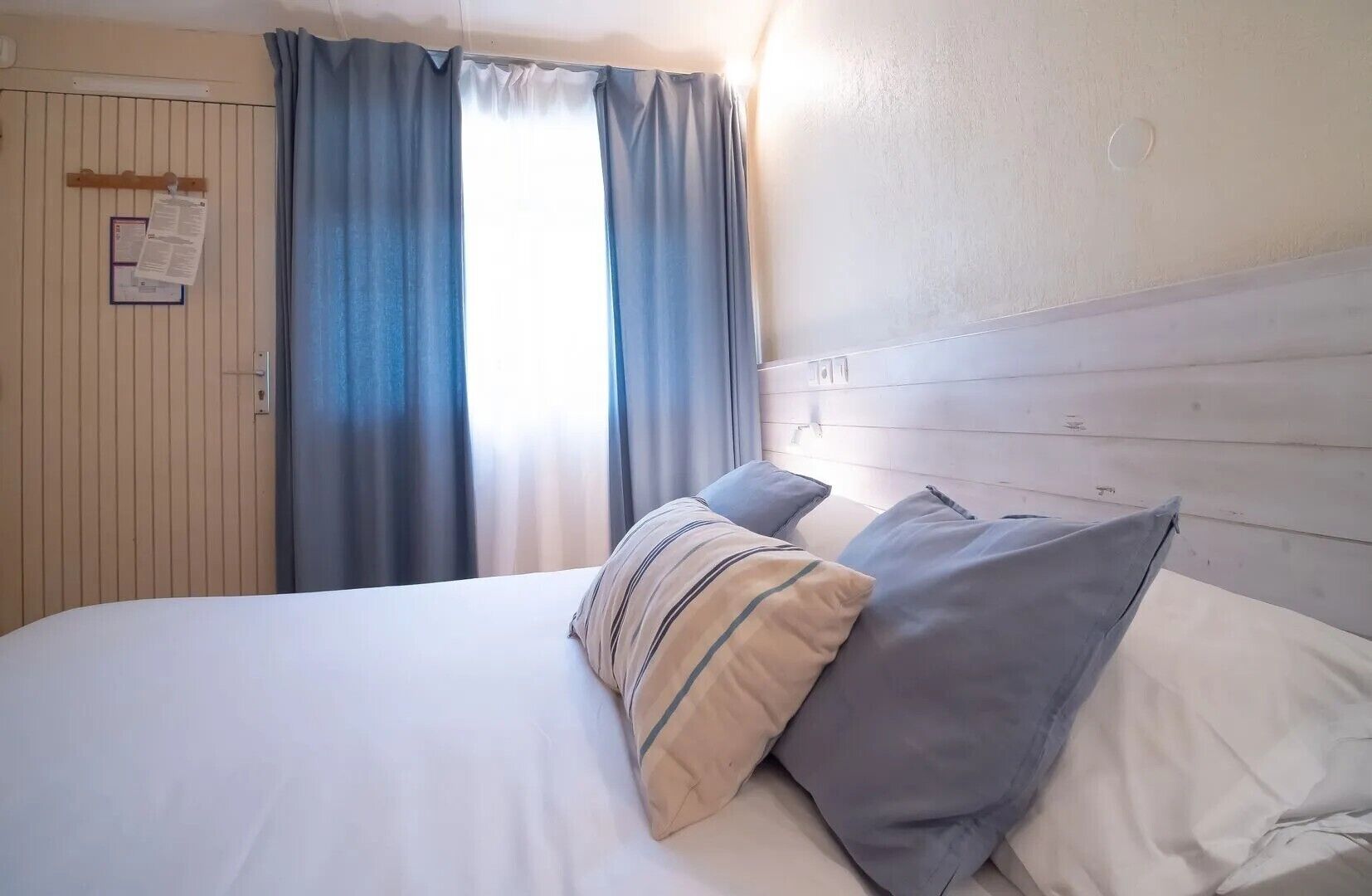 A bed with pillows, a wooden headboard, a window with curtains, and a wall-mounted notice.