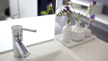 Un baño moderno con grifo cromado, lavamanos blanco y tres jarrones blancos con flores moradas.