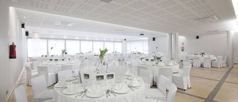 A large banquet hall with round tables set for events, white chairs, and a high ceiling with recessed lighting.