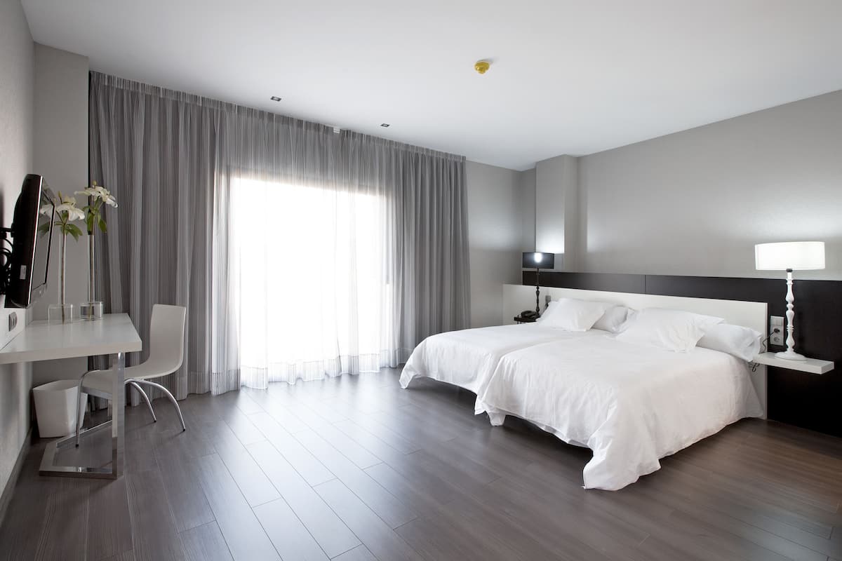 A modern hotel room with a large bed, a desk with a chair, and a large window with curtains.