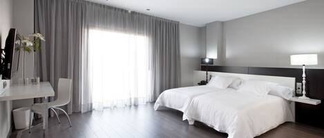 A modern hotel room with a large bed, a desk with a chair, and a large window with curtains.