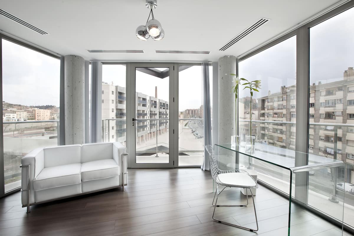A modern living room with a white sofa, a glass table, and a view of a cityscape through large windows.