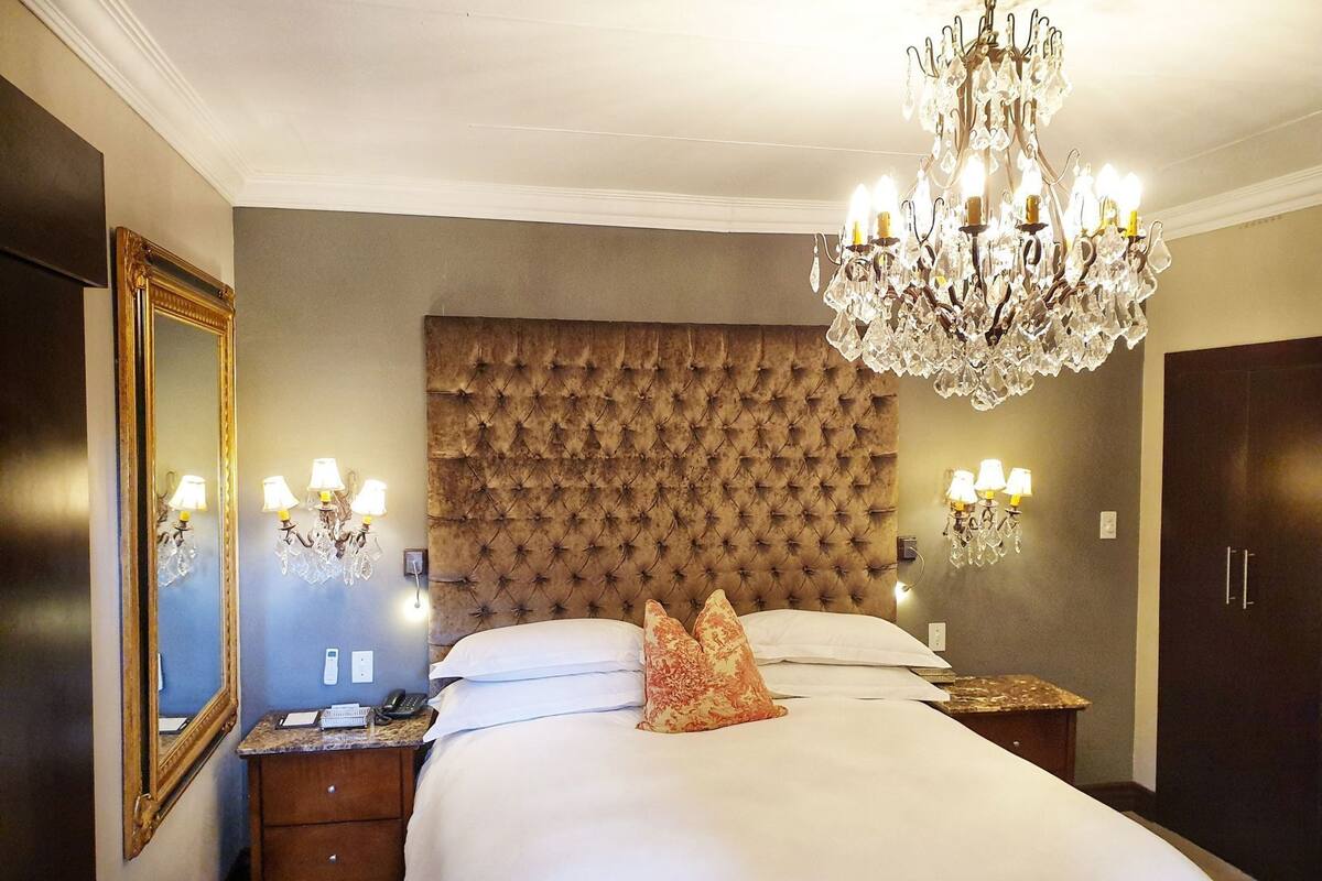 A bedroom with a tufted headboard, a large bed, a chandelier, bedside lamps, a mirror, and a wardrobe.