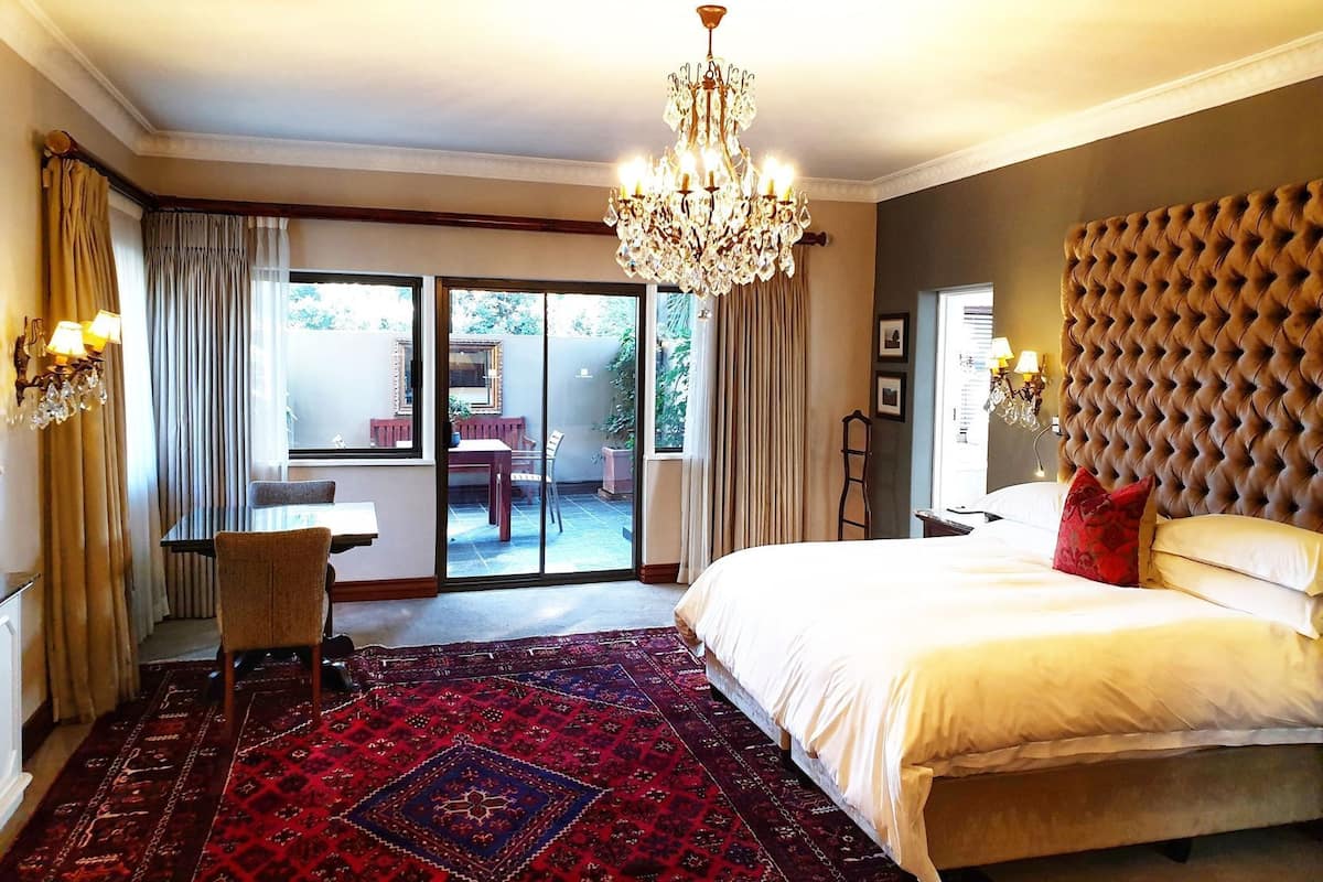 A bedroom with a large bed, a chandelier, a view to a patio with a table and chairs, and a red patterned carpet.
