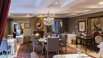 Une salle à manger dotée d’une cheminée, d’un lustre et de plusieurs tables dressées pour accueillir les convives.