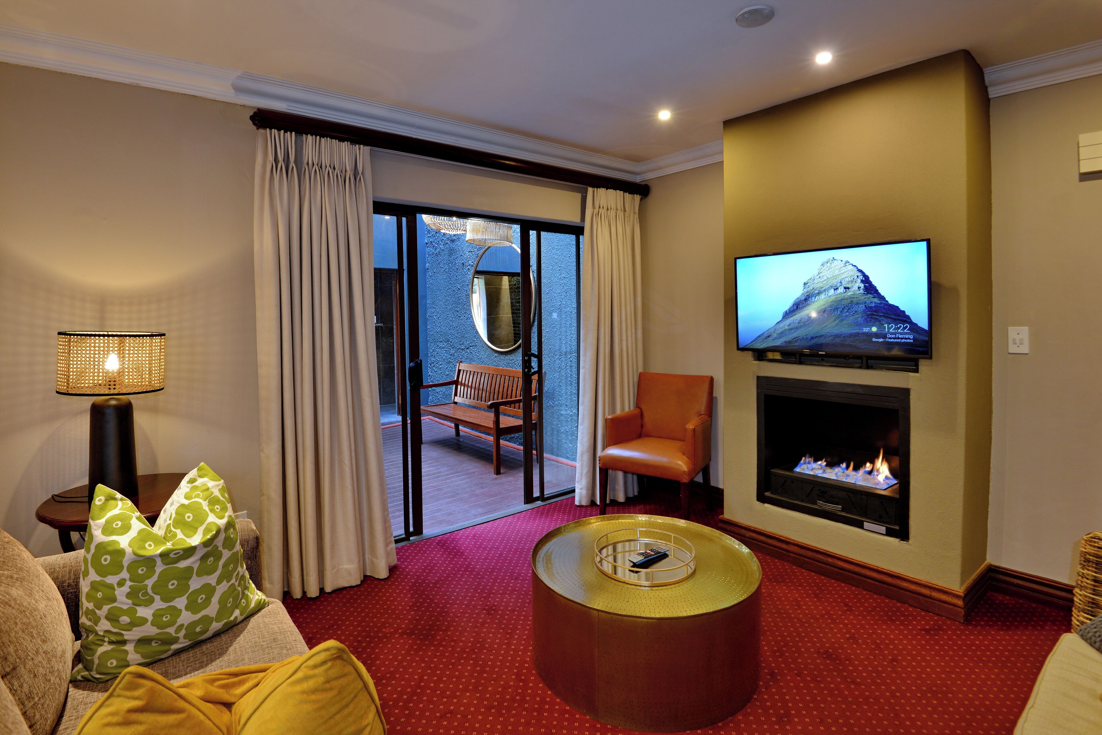 A hotel room with a fireplace, a television mounted on the wall, a sofa with patterned cushions, and a view through a glass door to an outdoor area with a bench and a chair.