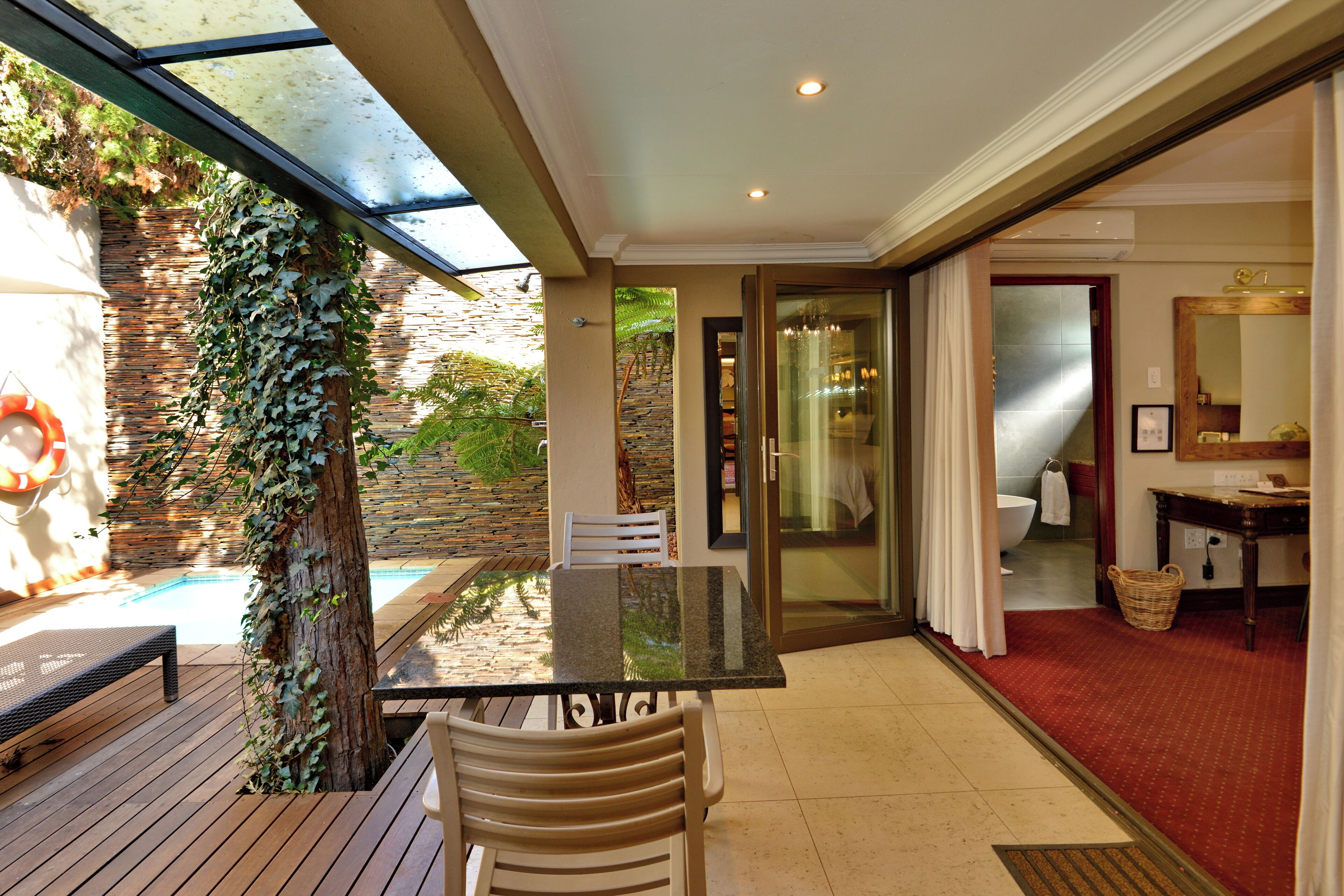 A sunlit patio with a pool, a table, and chairs, leading to a bathroom with a shower and a mirror.