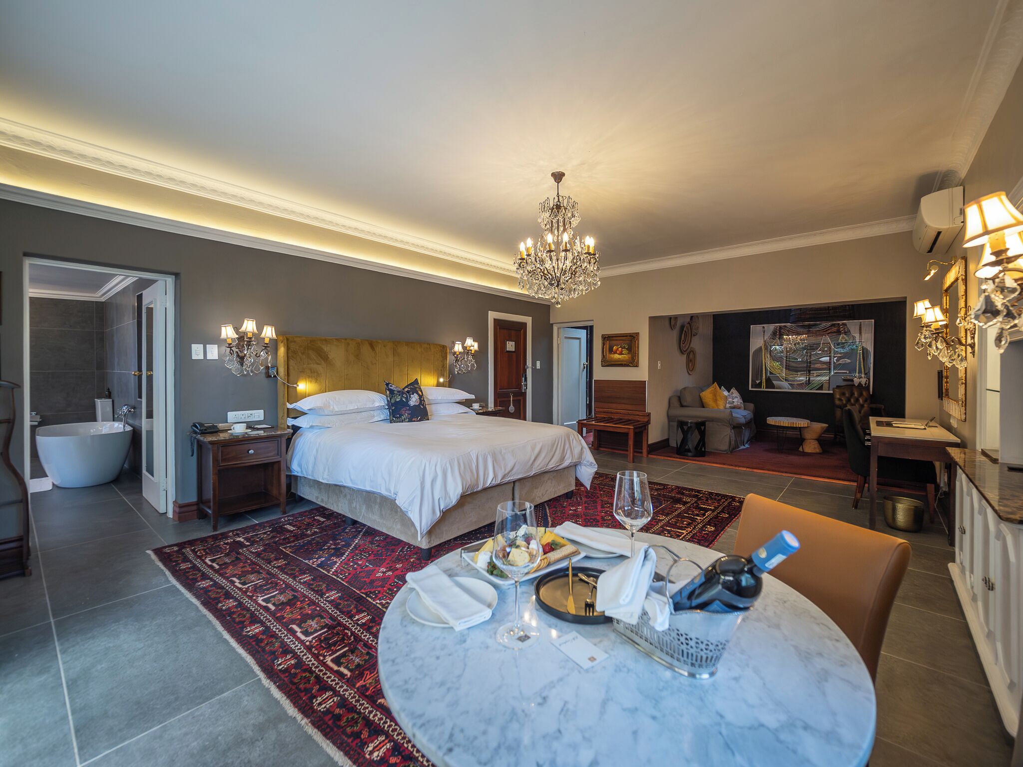 A hotel room with a large bed, a sitting area with a sofa and table, a bathroom with a bathtub, and a dining area with a table set for two.