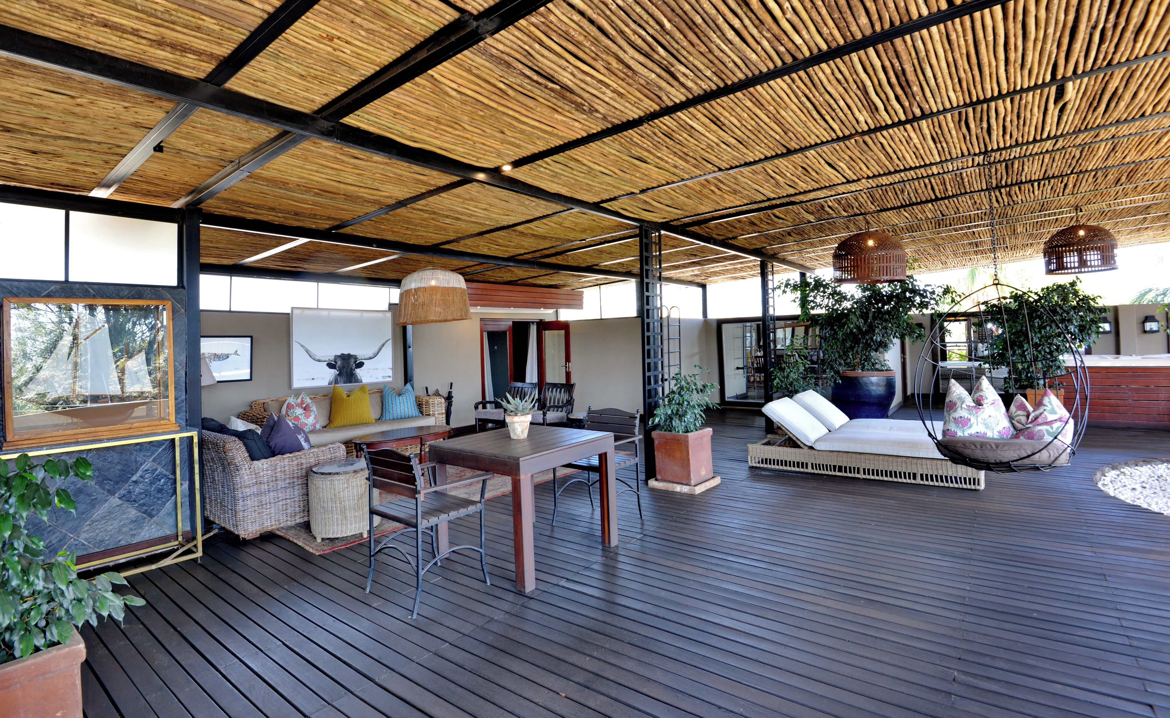 Un amplio espacio al aire libre con una terraza de madera, una mesa de comedor, sillas, un sofá y plantas colgantes.