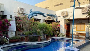 Una piscina con una escultura de delfín, rodeada de plantas en macetas y un área de patio con baldosas.