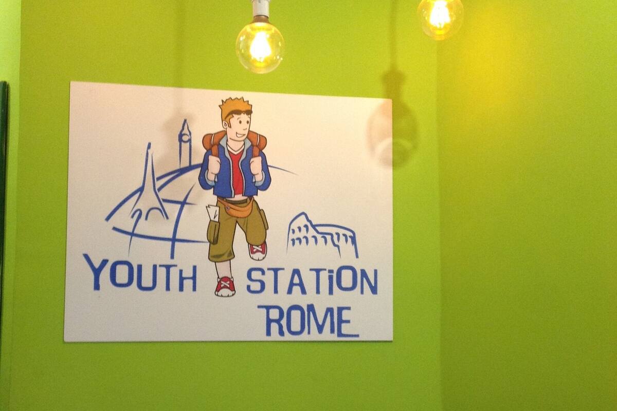 A wall with a sign reading "YOUTH STATION ROME" and a cartoon character. There are three hanging light bulbs above the sign.