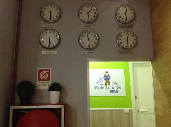 A wall with clocks labeled with city names like Cape Town, Sydney, Rio de Janeiro, Rome, New York, and Tokyo. Below are potted plants and a sign for a youth station.
