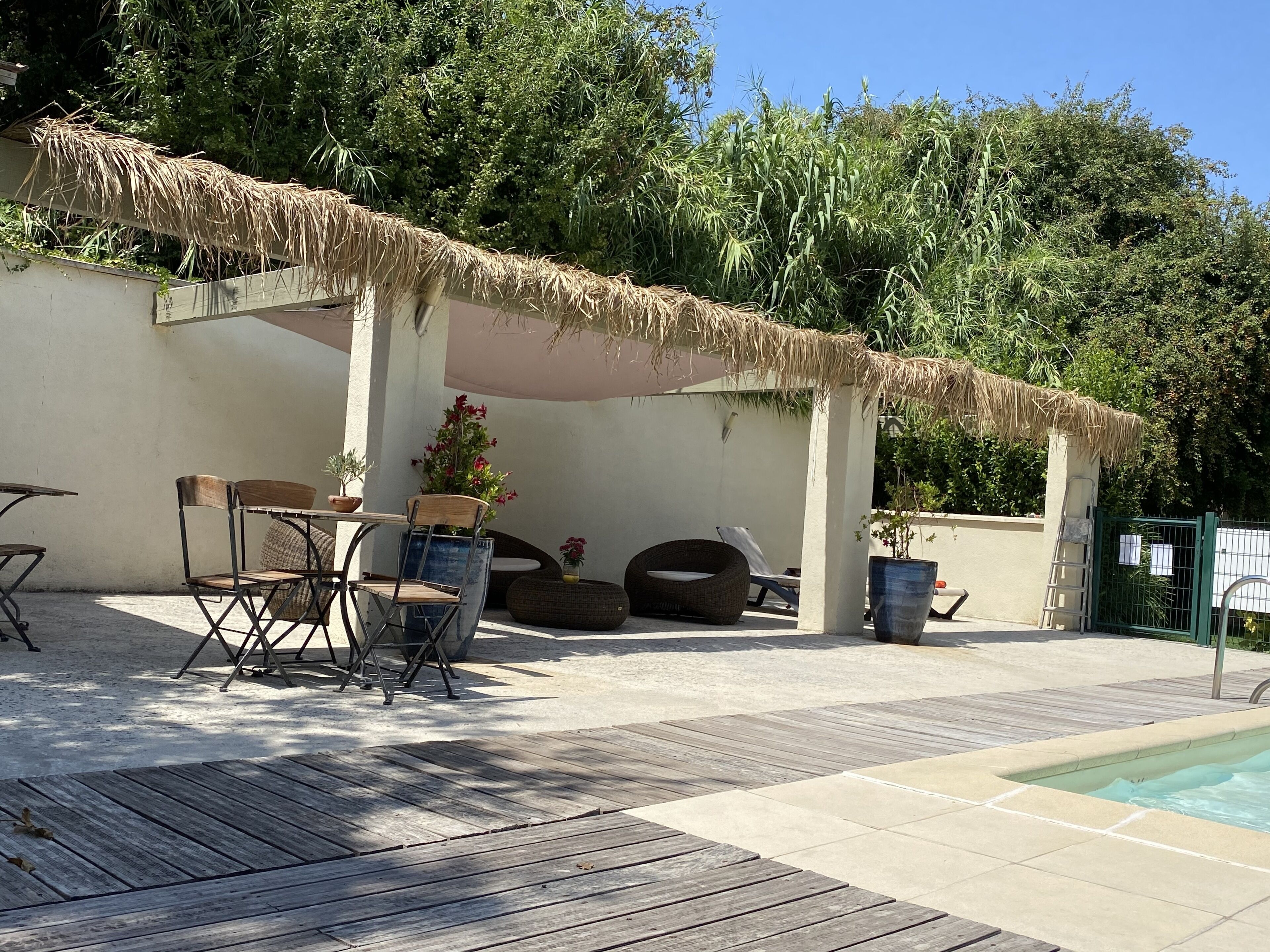 Ein Außenbereich mit Terrasse, Pool, Holzdeck, Sitzgelegenheiten und einem Reetdachgebäude.