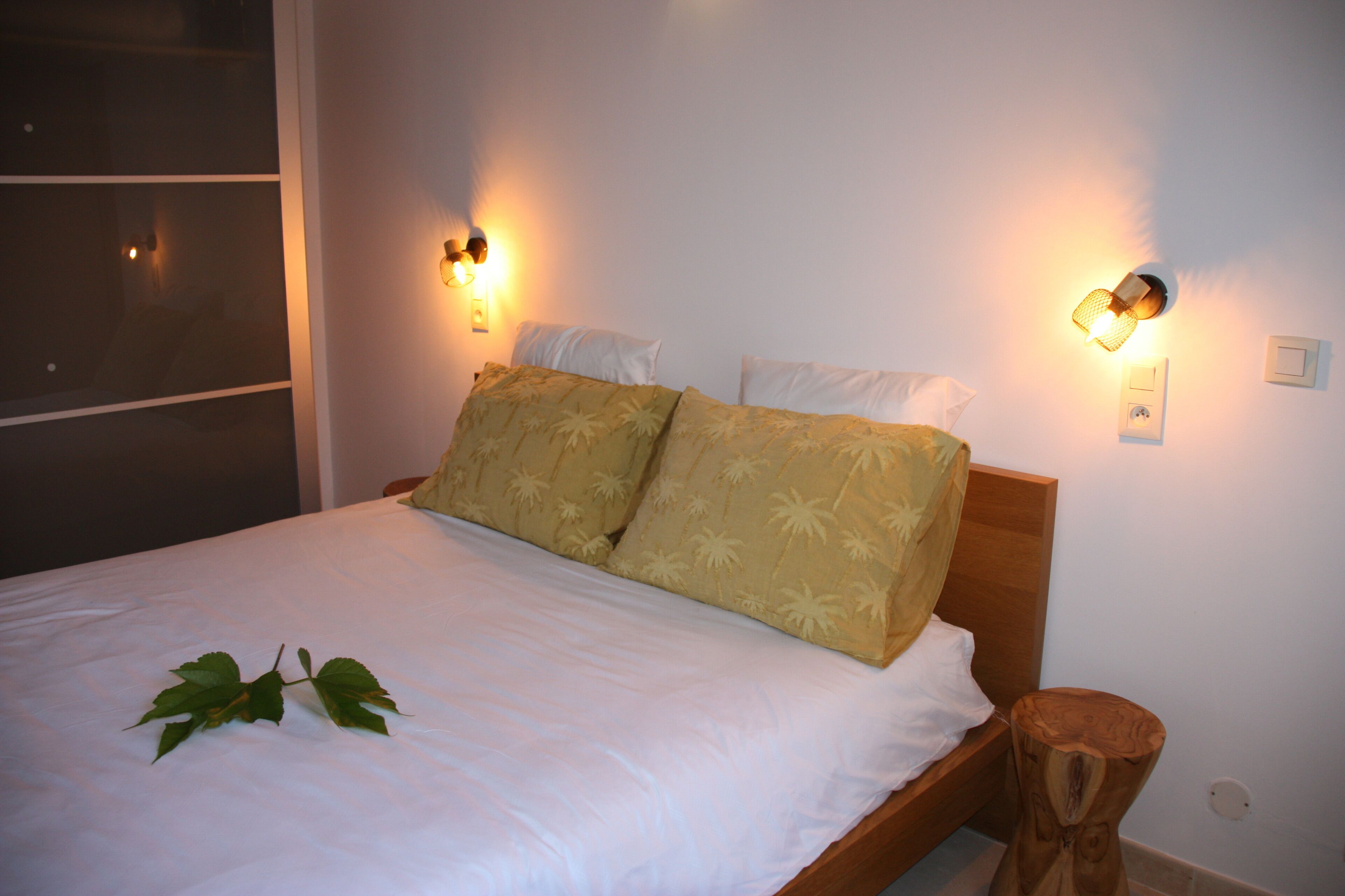 A neatly made bed with a white sheet and two pillows, a bedside table with a lamp, and a window with blinds.