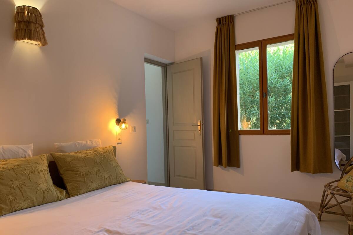 A bedroom with a bed, a wall-mounted lamp, a window with curtains, and a door.