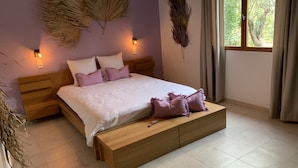 Een slaapkamer met een houten bed, wit beddengoed en paarse kussens. Er zijn wandlampen en een groot raam met gordijnen.