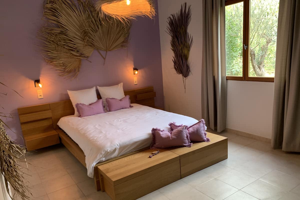 A bedroom with a wooden bed, white bedding, and purple pillows. There are wall-mounted lights and a large window with curtains.
