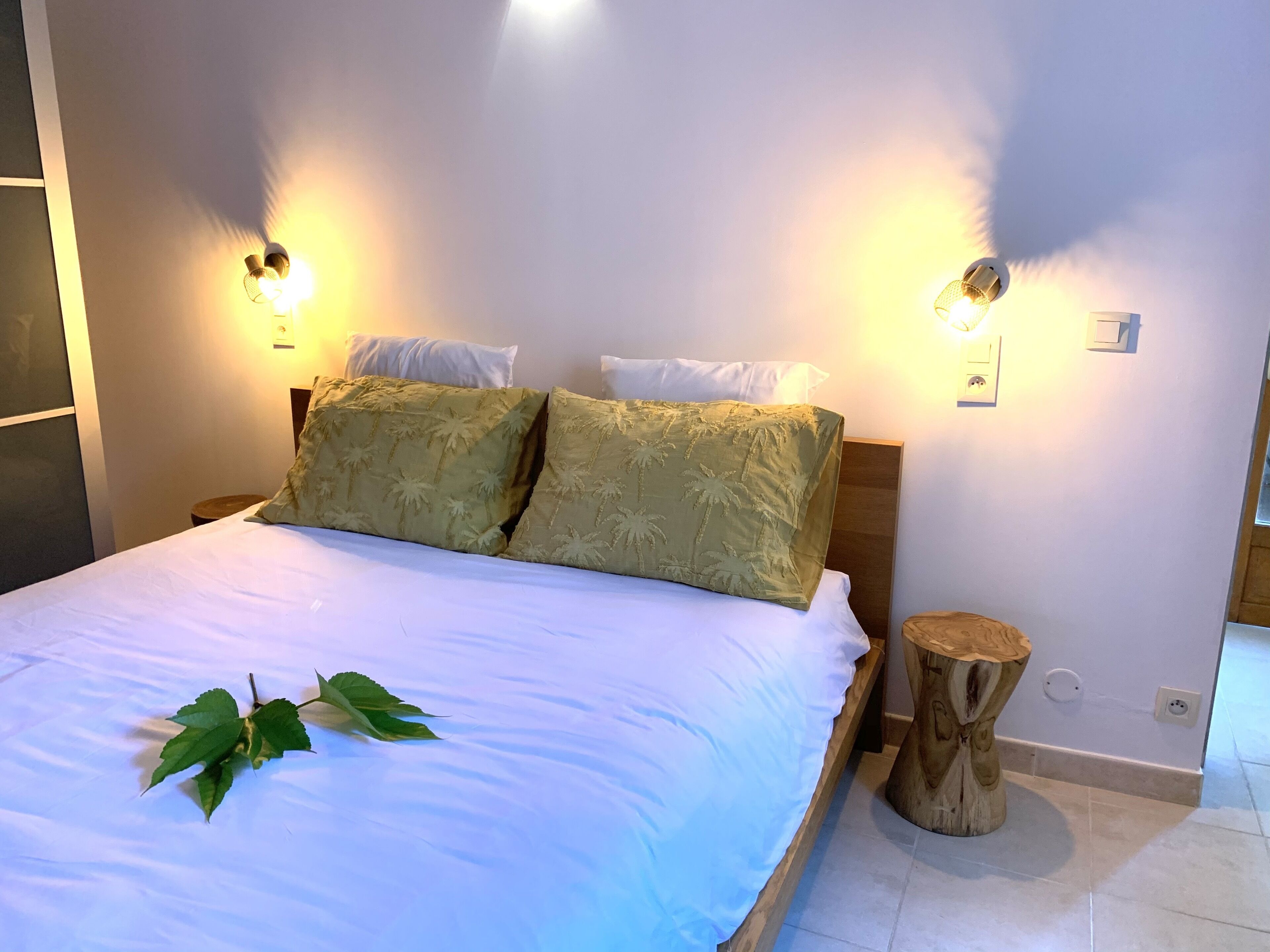 A neatly made bed with a white sheet and two green pillows, a bedside table with a wooden stool, and wall-mounted lights.