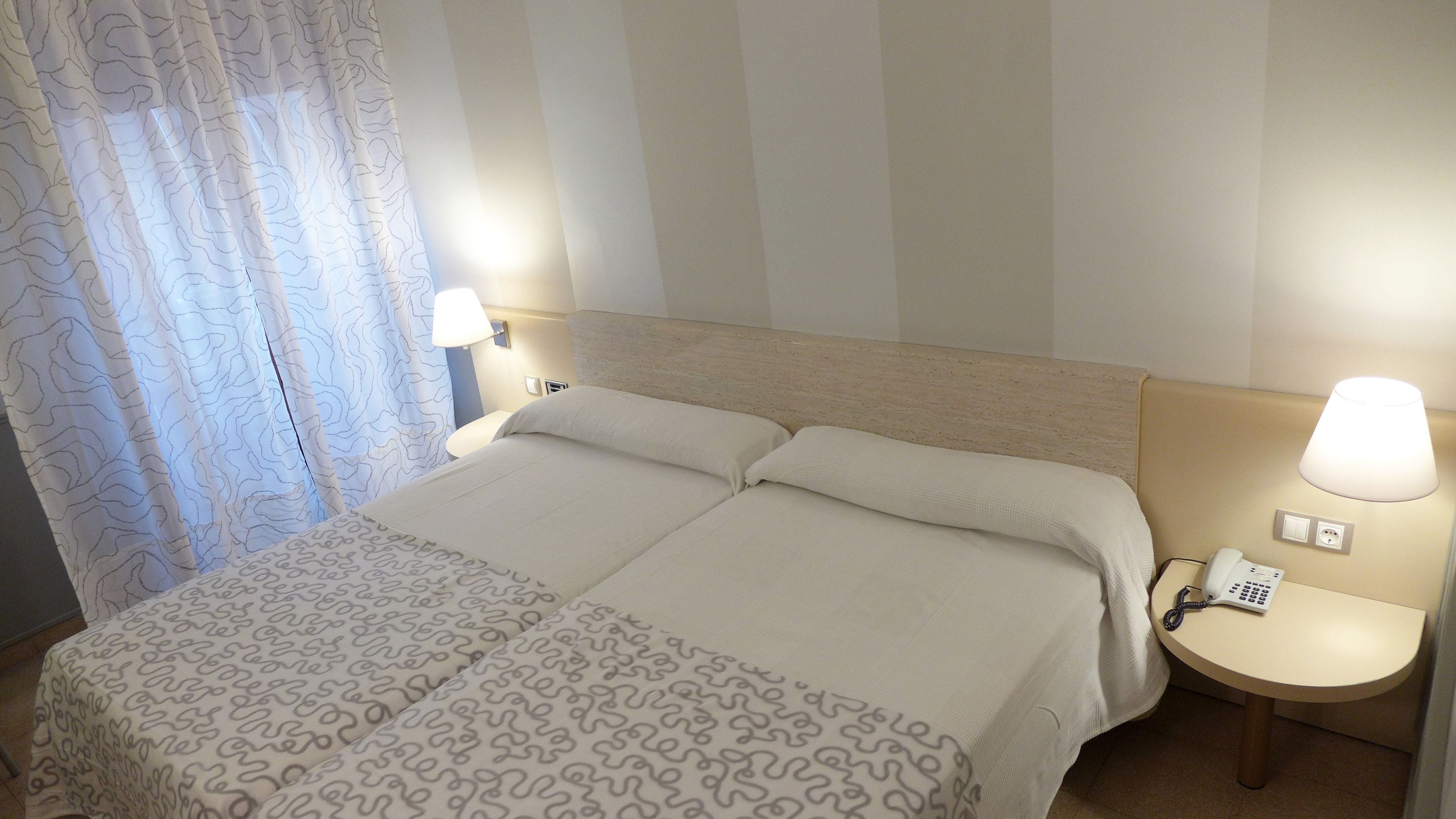 Una cama doble con cubrecama estampado, dos lámparas de noche, un teléfono sobre una mesita de noche y una cortina con diseño ondulado.