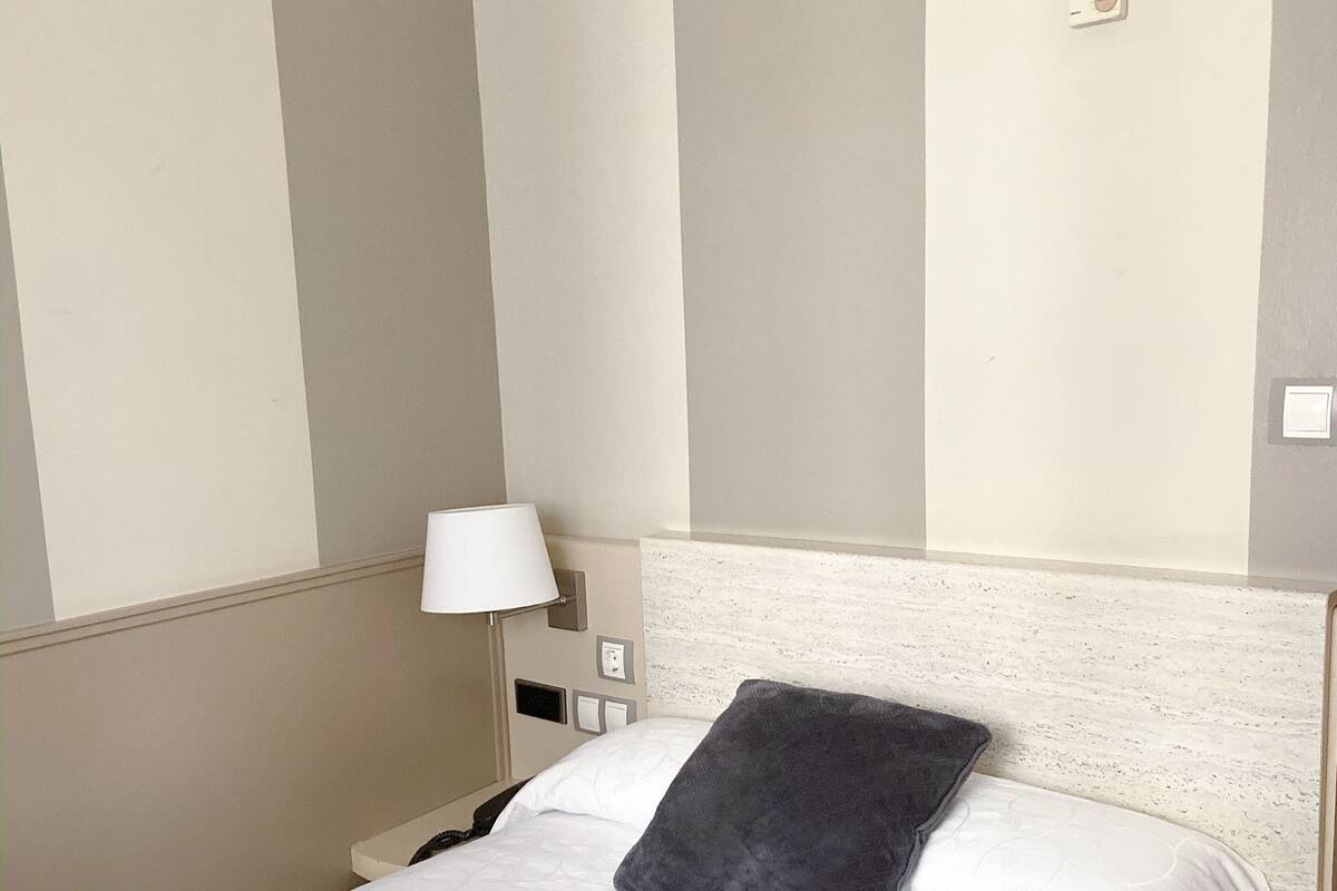 Quarto de hotel com uma cama, mesa de cabeceira, luminária e interruptor de luz na parede.