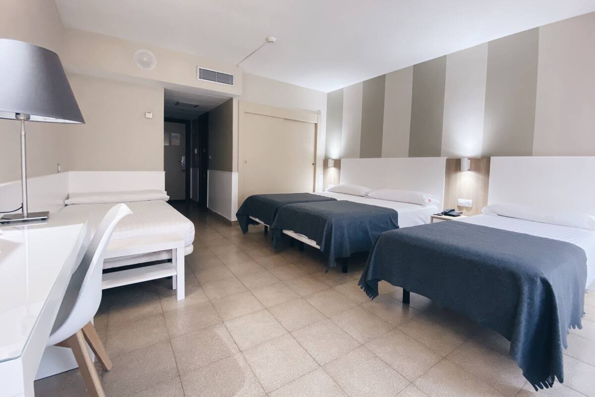 Quarto de hotel com duas camas de solteiro, uma escrivaninha, uma cadeira e mesa de cabeceira com abajur.