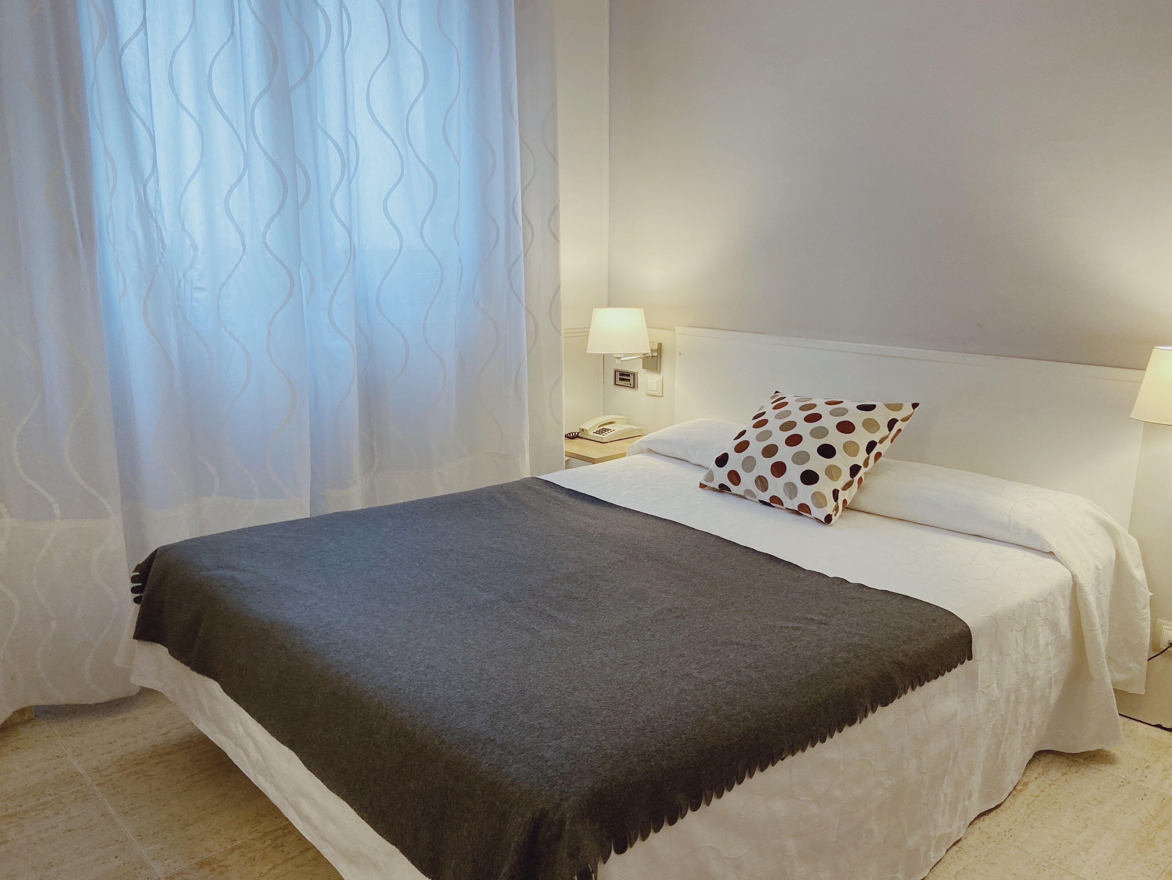 A neatly made bed with a grey blanket, white linens, and a polka-dotted pillow. There are two bedside lamps and a telephone on the nightstand.