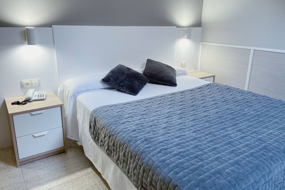 Cama bem arrumada com um edredom azul acolchoado, duas almofadas escuras e uma mesinha de cabeceira com um telefone branco.