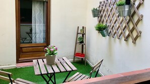 Un pequeño patio con una mesa y sillas de madera, una escalera de madera y una ventana con cortina.