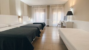 Habitación de hotel con tres camas individuales, un escritorio con silla y un ventanal con cortinas ligeras.
