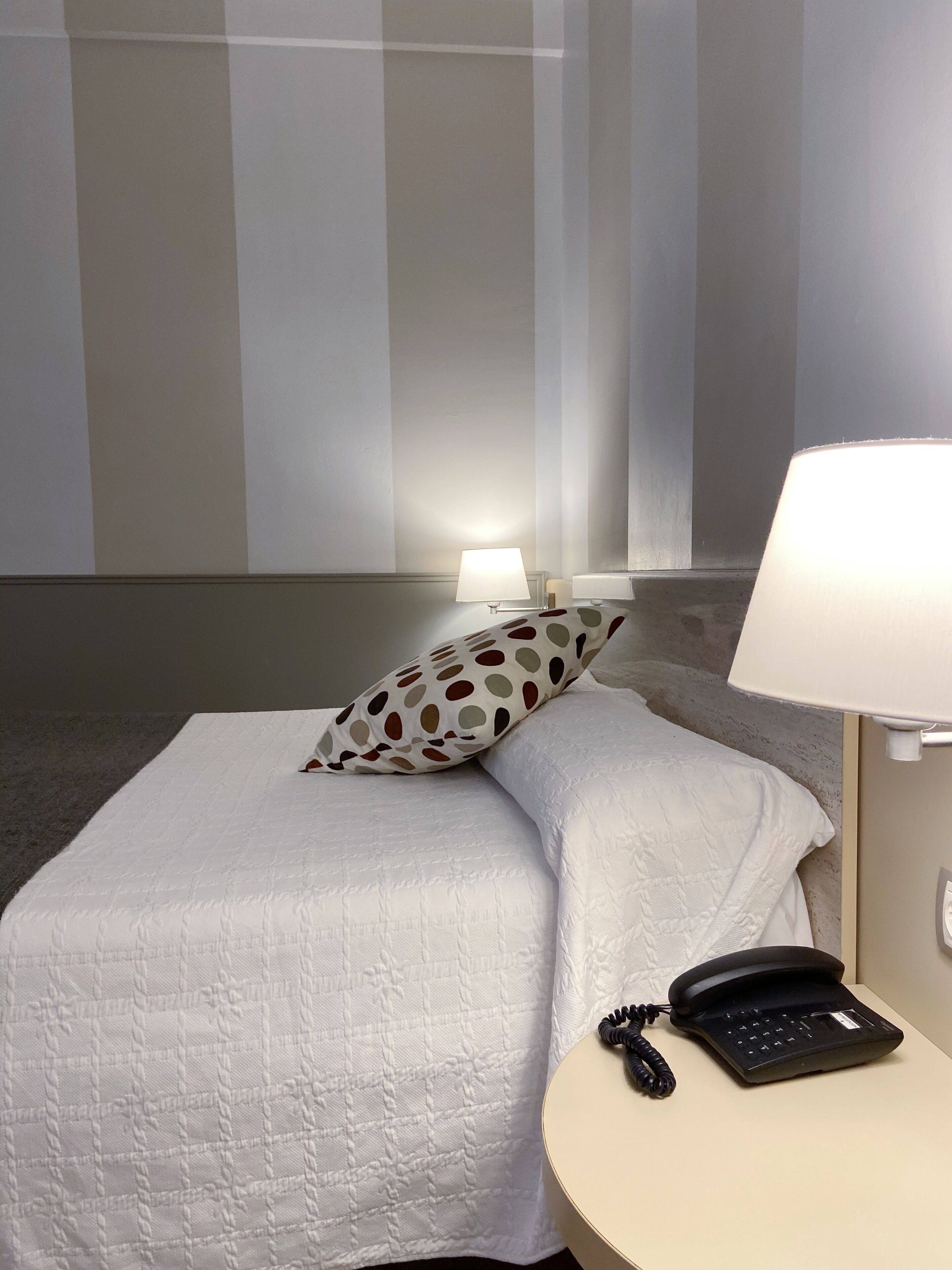 Habitación de hotel con una cama, almohadas, una lámpara de noche y un teléfono sobre una mesita.