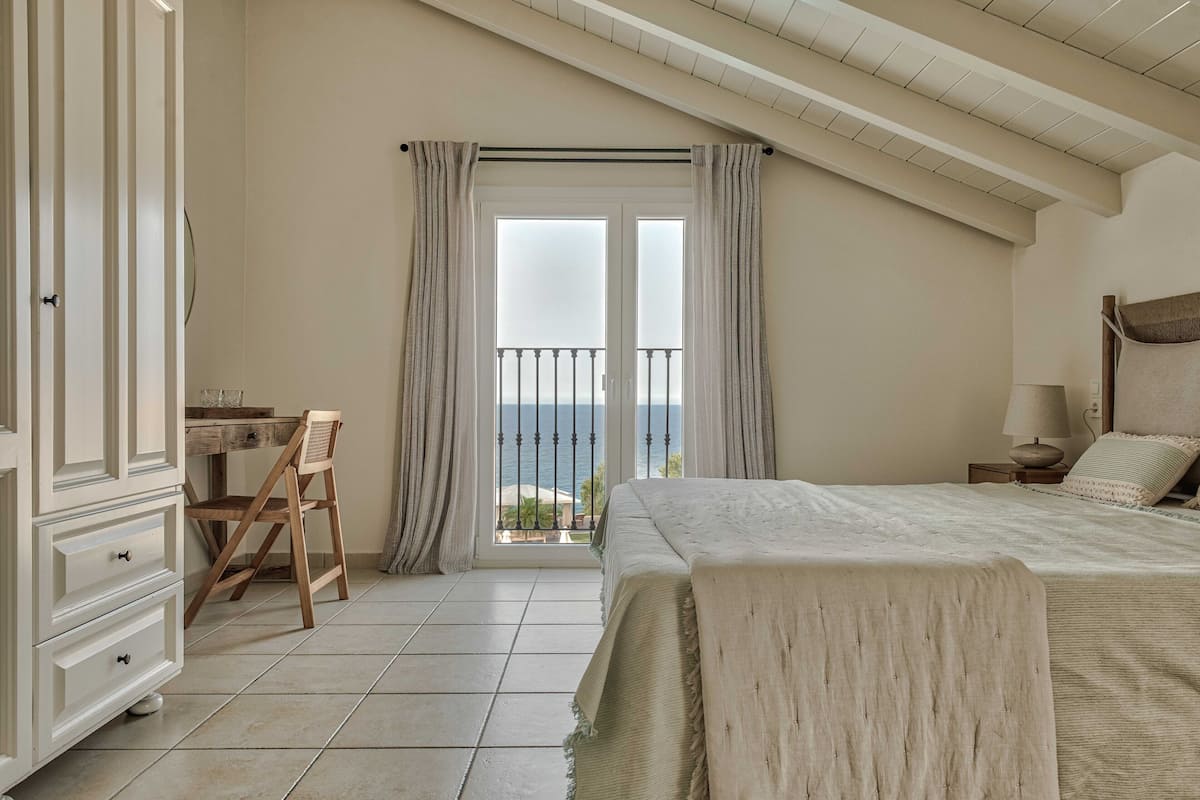 A bedroom with a bed, a desk, and a chair. There is a balcony with a view of the sea.